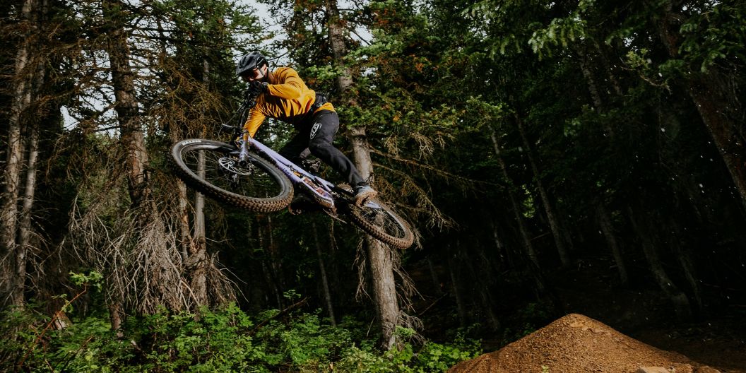 An MTB rider on an Ibis HD6 in fall apparel jumps a feature in an alpine tree forest.